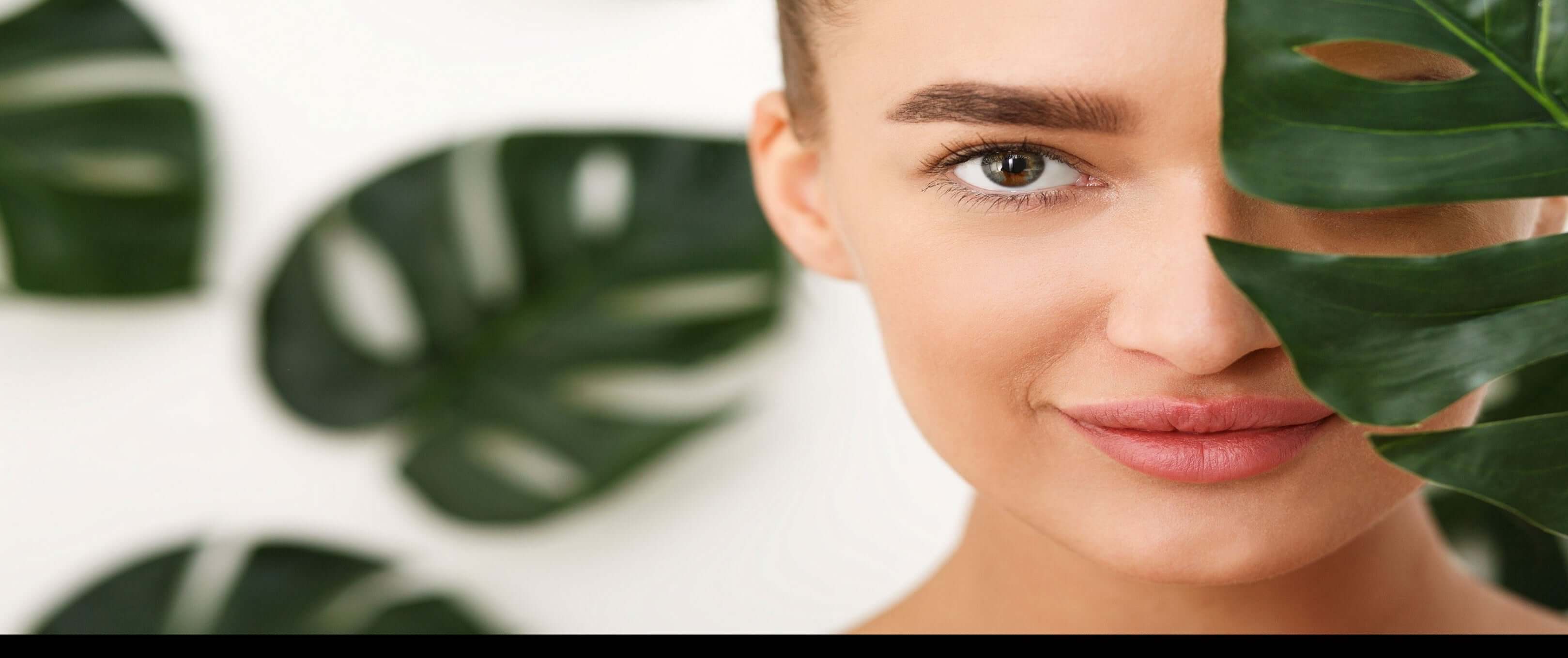 Close-up of a woman partially obscured by a leaf, showcasing her natural beauty and healthy skin.