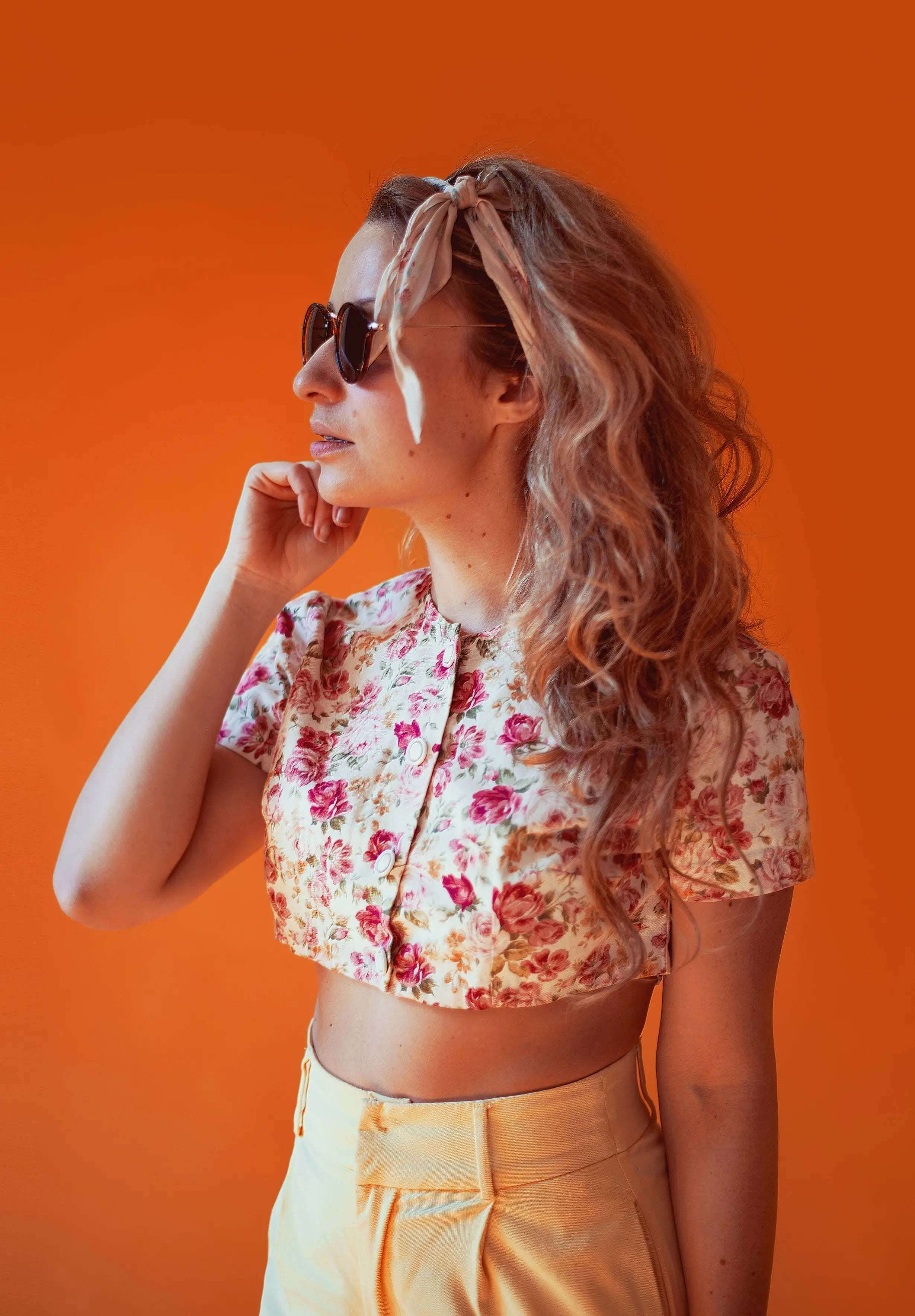 Woman wearing floral crop top and sunglasses against an orange background, showcasing a trendy summer style.