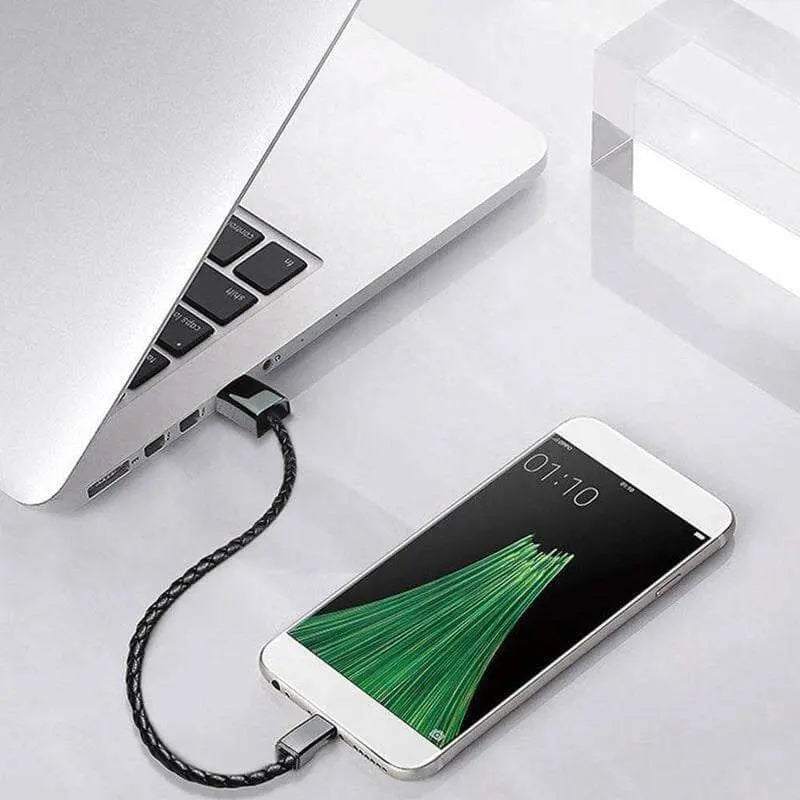 Black braided leather charge and sync cable bracelet connected between a laptop and smartphone for data transfer and charging.