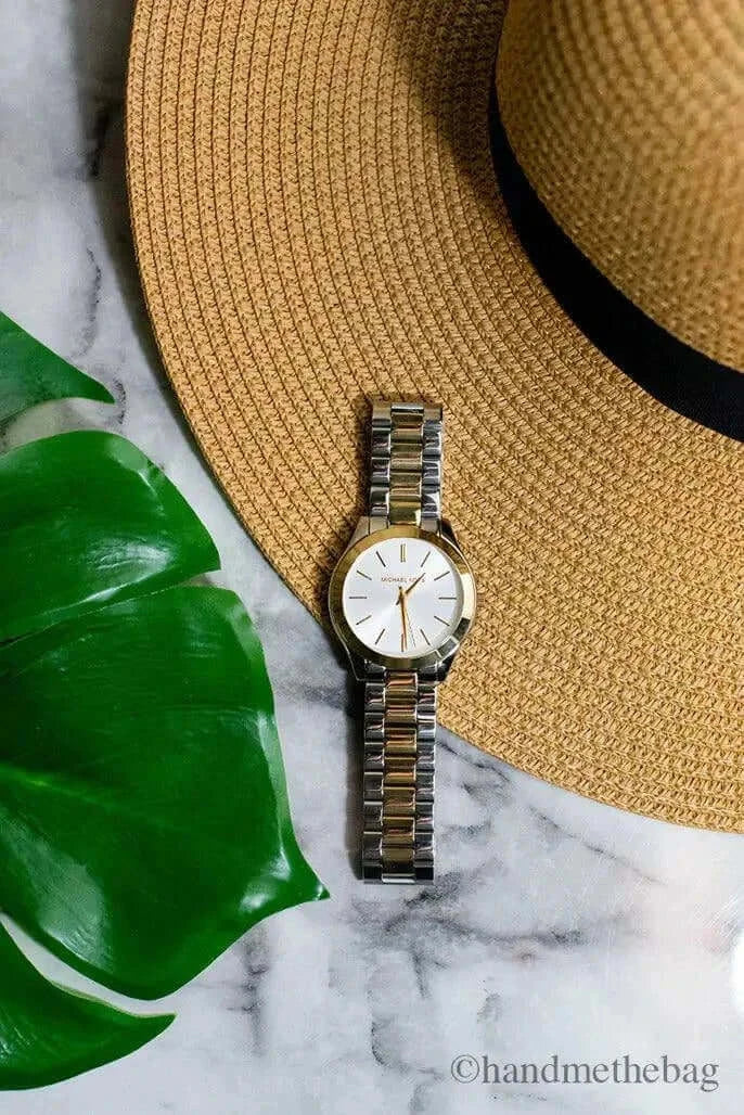 Michael Kors Slim Runway Gold Silver Toned Watch displayed with a straw hat and green leaf on marble surface.