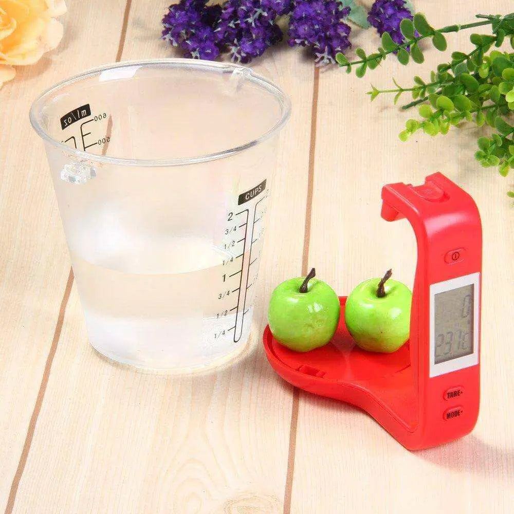 Digital measuring cup with LCD display, showing two apples on scale next to a clear measuring container on a wooden surface.